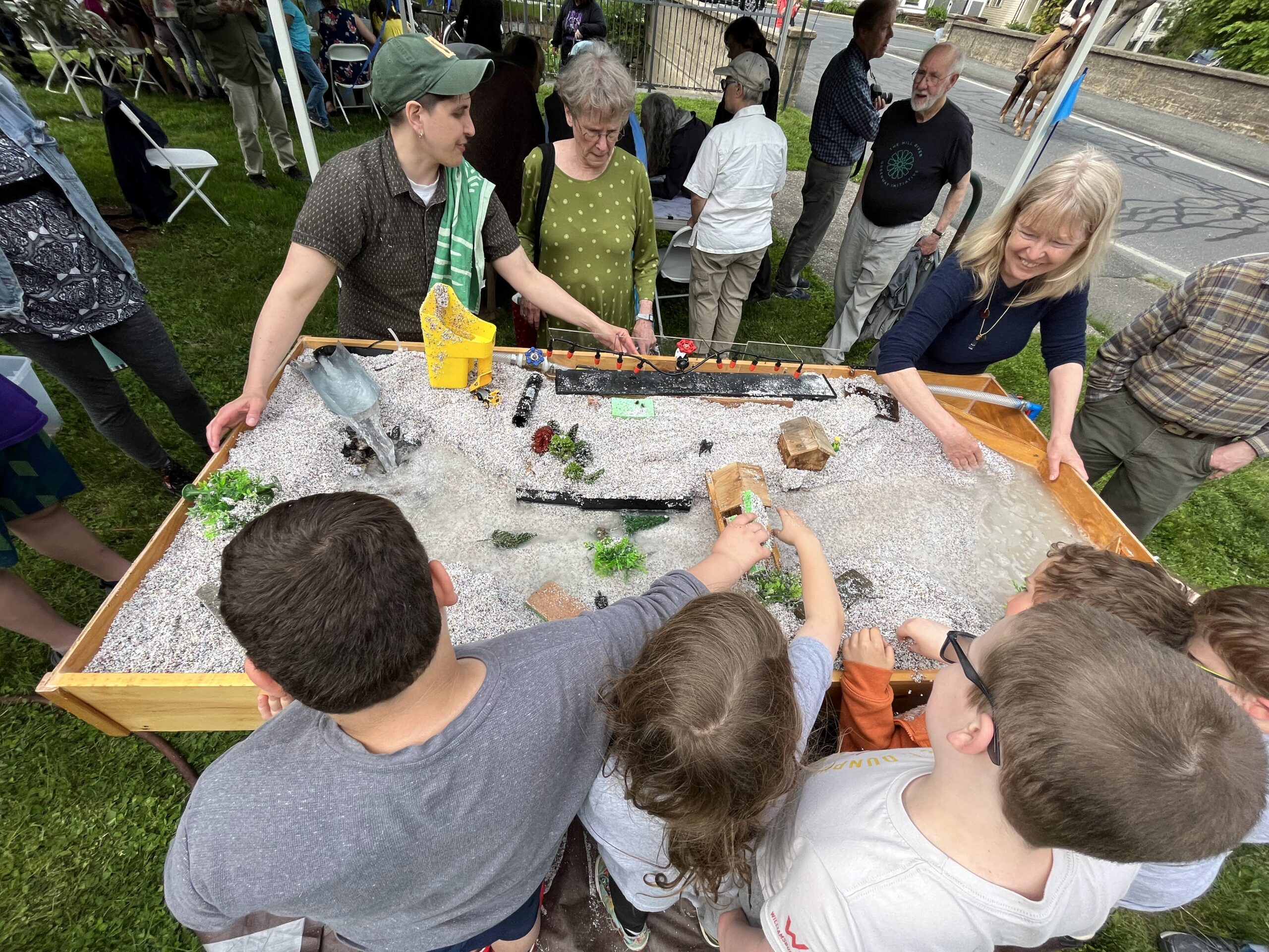 A group of people surrounds a table with a model of a river running through it. they are smiling and playing with scale model items like roads and houses.